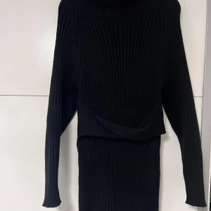 Svart ribbad stickad långklänning - Svart långklänning i ribbstickad design med polokrage och långa ärmar. Klänningen har en snygg omlottdetalj i midjan. Lite knottrig på vissa ställen.