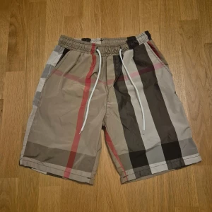 Beige-rutiga shorts med snörning i Burberry stil - Snygga shorts med klassiskt rutigt mönster i beige, svart, grått och röda detaljer. De har elastisk midja med vit snörning och två sidofickor. Perfekta för sommardagar och chill vibes. Materialet känns lätt och luftigt. Liten i storlek