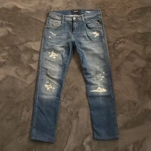 Replay Anbass slitna blå jeans - Säljer ett par Replay Anbass slim fit jeans i blå tvätt med slitna detaljer och snyggt distressed look. Klassisk femficksmodell med dragkedja och knapp. Jeansen har en smal passform och är perfekta för dig som gillar en modern streetstil. Ny pris 1500 