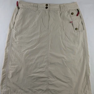 Beige långkjol med dragkedjor - Snygg beige långkjol från Tommy Hilfiger med coola detaljer som dragkedjor, knappar och röda band. Kjolen har praktiska fickor och justerbar nederkant med snörning. Perfekt för dig som gillar en avslappnad men ändå stilren look.Y2K style beige skirt  Size 42  Pockets with red zips  Elasticated pull cords at the bottom  70% cotton, 30% nylon  Waist approx 43cm  Length approx 63.5cm