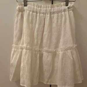 Vit volangkjol från NA-KD - Superfin vit kortkjol från NA-KD med volangdetalj och resår i midjan, vilket gör att den passar även som storlek 38. Kjolen har en lätt strukturerad yta och är perfekt för en fräsch och somrig look. Passformen är normal och materialet känns mjukt och luftigt.