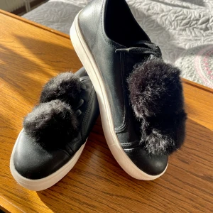 Svarta sneakers från Nelly - Svarta sneakers med vit sula och stora fluffiga svarta fuskpälsbollar på ovansidan. .