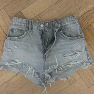 Gråa slitna jeansshorts - Säljer ett par gråa jeansshorts med hög midja och råa, fransiga kanter. Shortsen har slitningar och hål för en riktigt cool och avslappnad look. Perfekta för varma dagar när du vill ha en edgy stil.