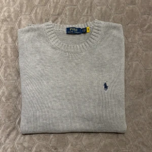 Ljusgrå stickad tröja från Polo Ralph Lauren - Snygg ljusgrå stickad tröja från Polo Ralph Lauren med klassisk rund halsringning och marinblå broderad logga på bröstet. Tröjan har ribbade muddar och är perfekt för lager-på-lager under höst och vinter.