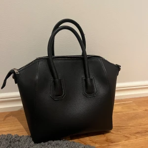 Svart handväska med dragkedja - Stilren svart handväska med två rejäla handtag och silverfärgad dragkedja. Väskan har rymligt huvudfack och innerficka med dragkedja. Perfekt för dig som vill ha en klassisk och snygg väska till vardagen.