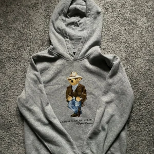 Grå Polo Bear hoodie från Ralph Lauren - Säljer en grå hoodie från Polo Ralph Lauren med ikoniska Polo Bear-trycket på bröstet. Tröjan har huva och är långärmad, tillverkad i mjuk bomullsmix. Perfekt för dig som gillar streetstyle och vill ha något unikt med stylish -vibe.