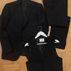 Svart kostym med väst från Tiger of Sweden - Klassisk svart kostym från bläck med kavaj, byxor och matchande väst. Stilren design med snygg passform och snygga detaljer. Perfekt för dig som vill ha en tidlös och elegant look. Materialet är mjukt och följsamt, vilket ger en bekväm känsla hela dagen. Kavaj 50 väst 46 byxor 148