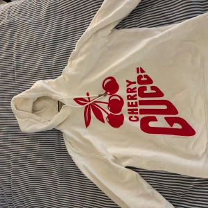 Vit hoodie från Gucci med körsbärstryck - Vit hoodie från Gucci i 100% bomull med stort rött körsbärstryck och texten 'CHERRY GUCCI' på framsidan. Klassisk huva med dragsko och långa ärmar. Snygg och unik streetstyle vibe, perfekt för dig som vill sticka ut. Givetvis är tröjan 100% äkta och är köpt i butik Frankrike. 