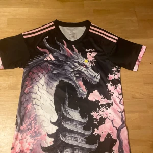 Japan Adidas fotbollströja med drake - Unik Japan fotbollströja från Adidas med sjukt snyggt drakmotiv, körsbärsblommor och pagoda. Svart bas med rosa detaljer och broderade loggor. Korta ärmar och lätt, sportigt material som andas. Perfekt för dig som vill sticka ut på planen eller gatan.