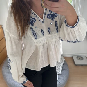 Vit blus  - Säljer en luftig vit blus med blå broderade detaljer och v-ringad hals. Blusen har långa, puffiga ärmar med broderier vid muddar och axlar. Materialet är lätt och känns som bomull, perfekt för vår och sommar. Passformen är loose och toppen har en bohemisk vibe. Den är i storlek xs men passar även S!