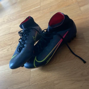 Nike svarta fotbollsskor med neon - Säljer ett par svarta Nike fotbollsskor med högt skaft och snörning. Skorna har neonfärgade detaljer i gult och rött, samt röda insidor. Sulan har fasta dobbar i grönt och svart, perfekt för gräsplan. Snygg och sportig design som sticker ut på planen. Pris kan diskuteras 