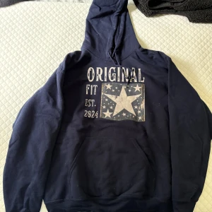 Mörkblå hoodie med stjärntryck - Mörkblå hoodie från Original Fit med stor huva och magficka. Framsidan har ett tryck med stjärnor och texten 'Original Fit Est. 2024'. Perfekt för chill dagar och streetstyle. Mjuk insida och klassisk passform.
