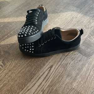 Louboutin skor - Svarta Louboutin skor i mocka med platt sula och rund tå. Skorna har snörning och är dekorerade med silvriga nitar över tån för en edgy look. Perfekta för dig som vill sticka ut med din stil. Insidan är ljusbeige och sulan är svart.