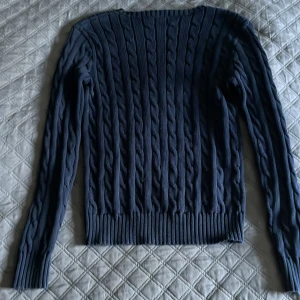 Mörkblå kabelstickad tröja Polo Ralph Lauren - Kabelstickad mörkblå tröja från Polo Ralph Lauren med klassisk rund hals och röd broderad logga på bröstet. Långärmad modell i mjuk bomull med ribbade muddar och nederkant. Perfekt för lager-på-lager och snygg till jeans.