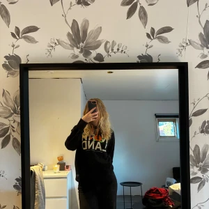 Norrlands hoodie - Svart hoodie från SQRTN Company med stor vit text 'NORRLAND' och ett tryck av hjortron i orange och grönt. Tröjan har huva med orange snören och en klassisk känguruficka framtill. Perfekt för dig som gillar streetwear och vill sticka ut. Tyvärr ett märke på N:et 