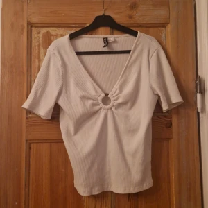 Vit ribbad topp med ringdetalj - Snygg vit ribbad topp från H&M med korta ärmar och djup v-ringning. Framtill finns en cool ringdetalj som ger toppen en unik look. Perfekt att styla med jeans eller kjol för en trendig outfit.