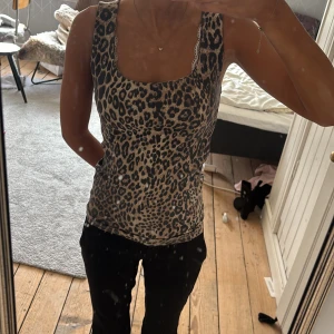 Leopardmönstrad ärmlös linnetopp - Snygg ärmlös linnetopp med leopardmönster och fyrkantig ringning. Toppen har en beige och svart färgskala och en liten spetsdetalj vid halsringningen. Perfekt för dig som vill sticka ut med ett djurmönster.