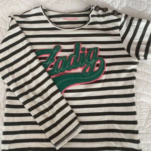 Randig långärmad tröja Zadig & Voltaire - Säljer en svartvit randig långärmad tröja från Zadig & Voltaire med grönt och rosa broderat 'Zadig'-tryck på bröstet. Tröjan har tre dekorativa knappar på axeln och är tillverkad i mjuk bomull. Perfekt för dig som gillar en cool och avslappnad stil.