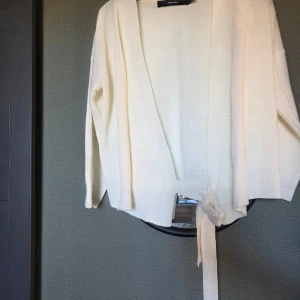 Vit omlottkofta från Vero Moda - Säljer en krämvit stickad omlottkofta från Vero Moda. Koftan har långa ärmar, breda ribbade kanter och knytband i midjan för en snygg passform. Perfekt att slänga över en topp när det är lite kyligt ute.