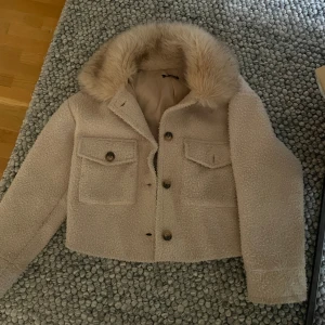 Beige teddyjacka med fuskpäls-krage - Säljer en beige teddyjacka med stor fluffig fuskpäls-krage. Jackan , har två stora bröstfickor med lock och knappar framtill. Perfekt för dig som vill ha en mysig och trendig look under höst och vinter. Jackan är inte nopprig utan är i teddymatrial de gör sig inte så rättvist på kameran. Sista bilden gör tyget mer rättvist