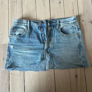 Replay jeanskjol med broderad detalj - Snygg ljusblå jeanskjol från Replay med rå kant nertill och slitna detaljer. Kjolen har klassiska fickor fram och bak, knappar framtill och en röd broderad patch med rosor bak i midjan. Perfekt för dig som gillar små tjolar!