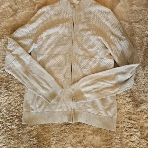 Ljusgrå kofta från Lindbergh White - Säljer en stilren ljusgrå kofta från Lindbergh White i storlek M. Koftan har dragkedja framtill, ribbade muddar och ett diskret strukturerat mönster på bröst och ärmar. Perfekt för lager-på-lager och enkel att matcha med det mesta.