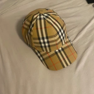 Burberry rutig keps i beige och svart - Snygg keps från Burberry med klassiskt rutigt mönster i beige, svart, vitt och röda detaljer. Justerbar rem med guldfärgad spänne bak och märkning på insidan. Perfekt för dig som vill ha en ikonisk streetstyle-accessoar.