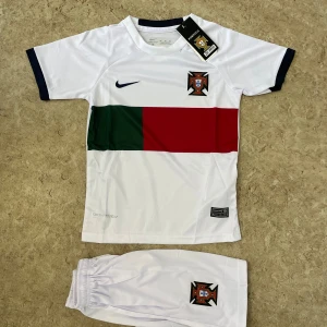 Portugal fotbollströja Nike vit - Säljer en vit Portugal fotbollströja från Nike med korta ärmar och svarta muddar. Framsidan har en bred grön och röd rand samt Portugal-emblem på bröstet. Materialet är lätt och andas, perfekt för träning eller match. Matchande vita shorts med emblem ingår.