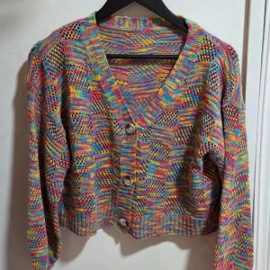 Färgglad stickad kofta med knappar, M - Superfin kofta i regnbågens alla färger med stickat, luftigt mönster och stora knappar framtill. Koftan har lång ärm, v-ringning och är perfekt för dig som vill sticka ut med en färgsprakande look. Passar lika bra till jeans som över en klänning.