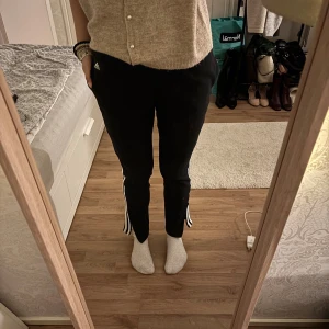 Svarta Adidas byxor med vita ränder - Svarta Adidas byxor med klassiska vita ränder längs sidorna och elastisk midja. Byxorna har en sportig look och är tillverkade i mjukt syntetmaterial, perfekta för chill eller träning. Adidas-logga framtill vid fickan.