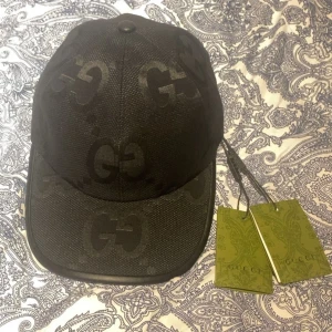 Gucci Keps  - Säljer en svart keps från Gucci med klassisk böjd skärm och stora GG-loggor ton-i-ton över hela kepsen. Kepsen är tillverkad i slitstark polyester och har justerbar rem baktill. Perfekt accessoar för att lyfta din streetwear-look.