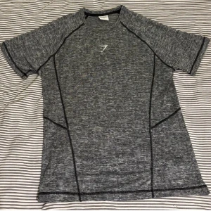 Grå träningst-shirt från Gymshark - Säljer en gråmelerad t-shirt från Gymshark med svart logga på bröstet. T-shirten har korta ärmar, svarta sömmar och är tillverkad i ett lätt och stretchigt syntetmaterial som passar perfekt för träning.