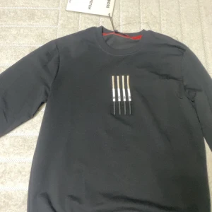 Svart långärmad tröja från Hugo Boss - Svart långärmad tröja från Hugo Boss med rund halsringning och broderad detalj på bröstet där det står BOSS i vitt, svart och guld. Röd insida på kragen med Hugo Boss-logga. Snygg och stilren design som sticker ut.