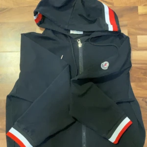 Svart vindjacka från Moncler med huva - Svart vindjacka från Moncler med huva och dragkedja framtill. Jackan har Moncler-logga på bröstet och snygga ränder i vitt och rött på ärmslut och huva. Perfekt för dig som vill ha en stilren och sportig look.