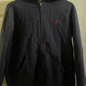 Mörkblå hoodie från Polo Ralph Lauren - Snygg mörkblå hoodie från Polo Ralph Lauren med dragkedja och huva. Klassisk röd broderad logga på bröstet och två fickor framtill. Perfekt för chill dagar eller när du vill ha en stilren streetlook.