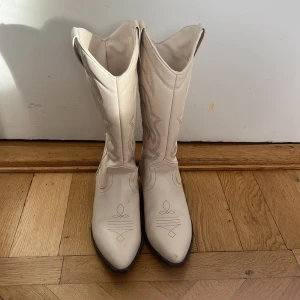 Vita cowboy boots med spetsig tå - Säljer ett par vita cowboy boots med snygga broderade detaljer och spetsig tå. Skorna har en låg klack och klassisk western-design. Tillverkade i skinnliknande material och har dragflikar upptill för enkel påtagning. Perfekta för att lyfta vilken outfit som helst.