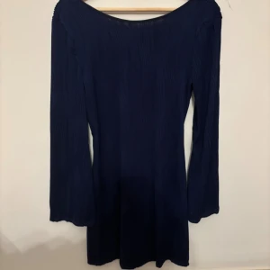 Mörkblå långärmad klänning med volangärm - Mörkblå kortklänning med lång ärm och snygga volangdetaljer vid ärmsluten. Klänningen har en lätt ribbad struktur och djup rygg med knytband. Sitter figurnära och har en stilren, enkel design som ändå sticker ut.