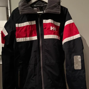 Helly Hansen jacka  - Seglarjacka från Helly Hansen med röda och vita ränder över bröst och armar. Jackan har hög krage i grå fleece, dragkedja framtill, huva med HH-logga och reflexdetaljer på ärmar och huva. Perfekt för blåsiga dagar och outdoor.