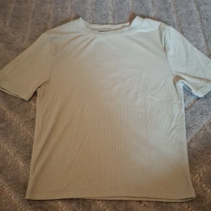 Grön ish ribbad t-shirt - Snygg grön ish t-shirt med ribbad struktur och klassisk rund hals. T-shirten har korta ärmar och är gjord i ett mjukt material som känns skönt mot huden. Perfekt för dig som gillar stilrena och enkla plagg med lite extra detalj.