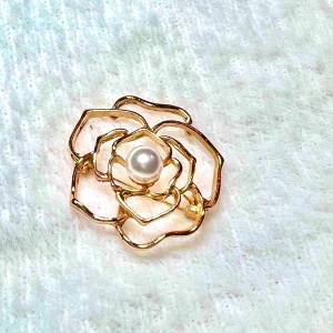 Guldfärgad blomformad brosch med pärla - Unik brosch i form av en blomma med guldfärgad metall och en vit pärla i mitten. Den öppna designen ger ett elegant och modernt intryck, perfekt för att lyfta vilken outfit som helst. Fästes enkelt med nål på baksidan.