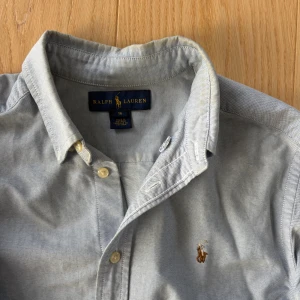 Ljusblå skjorta från Ralph Lauren - Klassisk ljusblå skjorta från Ralph Lauren med broderad logga på bröstet. Skjortan har lång ärm, knappar framtill och button-down krage. Tillverkad i mjuk bomull som känns skön mot huden. Perfekt för en clean och stilren look.