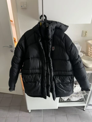 Svart dunjacka från Fjällräven S - Säljer en svart dunjacka från Fjällräven i storlek S. Jackan har huva, stora fickor framtill och dragkedja med gul detalj. Perfekt för kalla dagar och riktigt skön att bära. Klassisk Fjällräven-logga på bröstet.