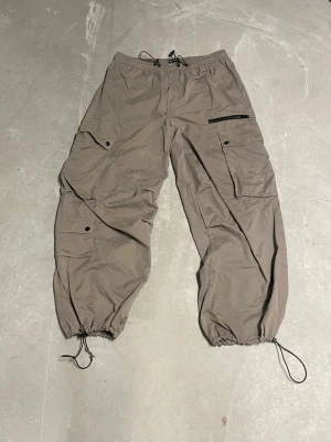 Beige cargopants med snörning - Säljer ett par beige cargopants med flera fickor och svarta dragkedjedetaljer. Byxorna har snörning i midjan och vid bensluten för att justera passformen. Materialet är lätt och syntetiskt, perfekt för en chill och avslappnad stil. Storleken är L/XL men psssae mig som vanligtvis har M eller S