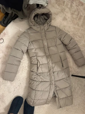 Beige lång pufferjacka med huva - Säljer en beige lång pufferjacka med huva och dragkedja. Jackan har quiltad design och två sidofickor med dragkedja. Perfekt för kalla dagar och har en stilren look som funkar till många outfits.