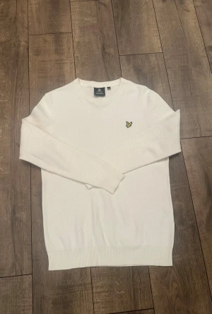 Vit stickad tröja från Lyle & Scott - Vit stickad tröja från Lyle & Scott i storlek M. Klassisk rund hals och långärmad modell med ribbade muddar. På bröstet sitter den ikoniska gula örn-loggan. Materialet är mjukt och har en diskret struktur som ger en schysst look.