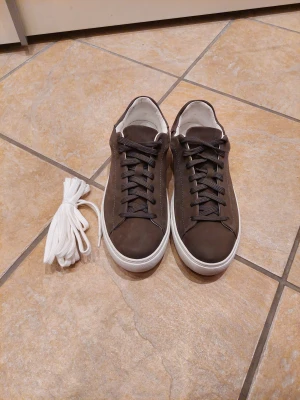 Bruna/Grå  sneakers i mocka med vit sula - Snygga bruna sneakers från Bruun & Stengade i mjuk mocka med vit, platt sula och svarta snören. Skorna har en stilren och minimalistisk design med rund tå och extra vita skosnören medföljer. Perfekta för dig som gillar en clean och modern look.