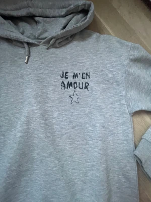 Grå hoodie från Gina Tricot med tryck - Säljer en grå hoodie från Gina Tricot med svart texttryck 'JE M'EN AMOUR' och en liten stjärna på bröstet. Tröjan har huva med snörning, långa ärmar och ribbade muddar. Perfekt för chill dagar och enkel att matcha med jeans eller mjukis.