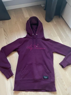 Mörklila Peak Performance hoodie XS - Snygg mörklila hoodie från Peak Performance med broderad logga i rosa på bröstet. Tröjan har huva med snörning, magficka och ribbade muddar. Materialet är mjuk bomull med insida i fleece för extra komfort. Perfekt för chill eller när du vill vara extra cozy.