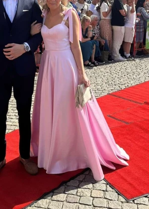 Ljusrosa långklänning med rosettband - Ljusrosa långklänning i glansigt satinliknande material med breda axelband som knyts i rosetter. Klänningen har markerad byst, v-ringad urringning och en svepande kjol med mycket vidd. Perfekt för bal. Har några fläckar längst ner men som inte syns så mycket.