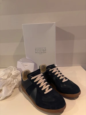 Maison Margiela gats  - Maison Margiela GAT sneakers i mörkblå mocka och skinn med beige detaljer och snörning. Klassisk rund tå och platt gummisula i brunt. Snygg minimalistisk design med vit insida och diskret branding på sulan. Perfekt för dig som gillar stilrena och exklusiva sneakers. Fråga på💫💫
